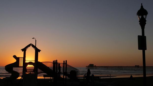 Oceanside, California USA - 11 Feb 2020: Silhouette of waterfront playground near pier. Children playing with parents, sunset ocean beach. Family with kids, beachfront recreation area, pacific coast.