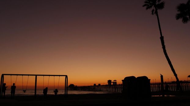 Oceanside, California USA - 8 Feb 2020: Silhouette of waterfront playground near pier. Children playing with parents, sunset ocean beach. Family with kids, beachfront recreation area, pacific coast.