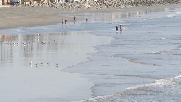 Oceanside, California USA - 11 Feb 2020: Local people walking strolling, pacific ocean coast, beach from pier. Sea water waves tide, shore sand. Beachfront vacations resort. Waterfront promenade.