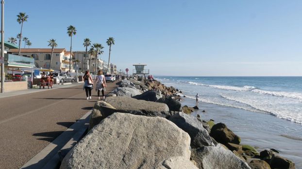 Oceanside, California USA - 16 Feb 2020: People walking strolling on waterfront sea promenade, beachfront boardwalk near pier. Vacations ocean beach resort near Los Angeles. Surrey bike bicycle riding