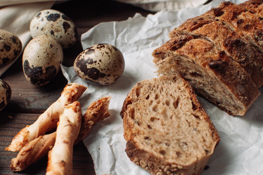 set dark yeast-free buckwheat bread in a cut lies on parchment, next to quail eggs and Italian grissini on a linen tablecloth on a wooden table in a rustic style. Breakfast cooking concept.