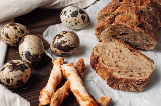 set dark yeast-free buckwheat bread in a cut lies on parchment, next to quail eggs and Italian grissini on a linen tablecloth on a wooden table in a rustic style. Breakfast cooking concept.