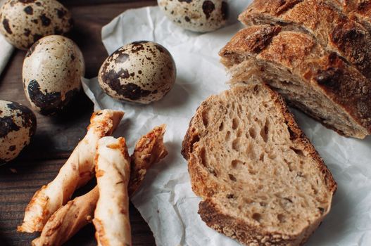 set dark yeast-free buckwheat bread in a cut lies on parchment, next to quail eggs and Italian grissini on a linen tablecloth on a wooden table in a rustic style. Breakfast cooking concept.