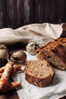 set dark yeast-free buckwheat bread in a cut lies on parchment, next to quail eggs and Italian grissini on a linen tablecloth on a wooden table in a rustic style. Breakfast cooking concept.