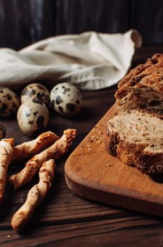 set dark yeast free buckwheat bread in a cut lies on a chopping wooden board, next to quail eggs and Italian grissini on a linen tablecloth on a wooden table in rustic style.Breakfast cooking concept.