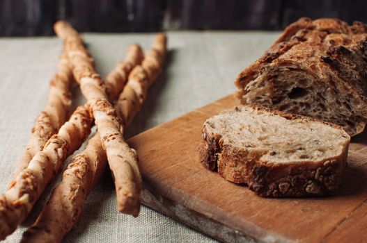 dark yeast-free buckwheat bread in a cut lies on a cutting wooden board on a wooden table, next to it is Italian grissini on a linen tablecloth in a rustic style. Breakfast cooking concept.