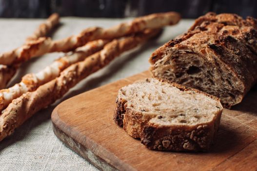 dark yeast-free buckwheat bread in a cut lies on a cutting wooden board on a wooden table, next to it is Italian grissini on a linen tablecloth in a rustic style. Breakfast cooking concept.