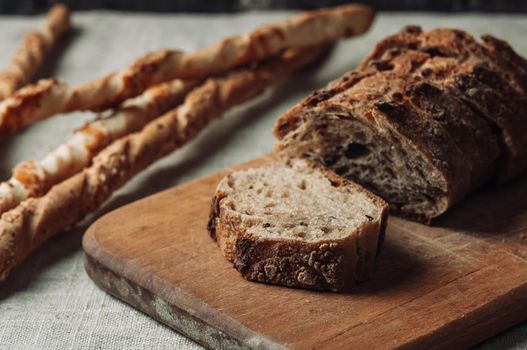 dark yeast-free buckwheat bread in a cut lies on a cutting wooden board on a wooden table next to italian grissini on a linen tablecloth in rustic style. Selective focus. Concept of making breakfast.