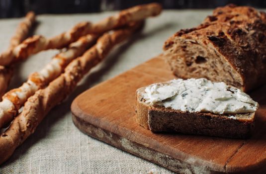 dark bread is spread with cottage cheese with herbs in a cut on a wooden board, next to a grissini on a linen tablecloth on wooden table in a rustic style. Snack and breakfast concept.Selective focus.