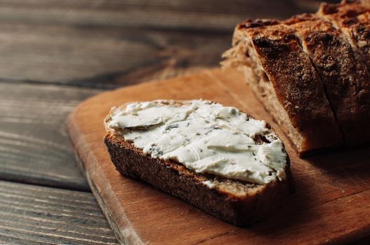 dark bread is spread with cottage cheese with herbs in a cut on a wooden board on a wooden table in a rustic style. Snack and breakfast concept. Selective focus.