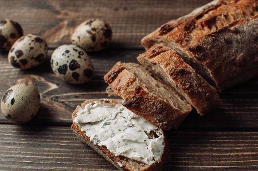 dark buckwheat bread with sisam is spread with cottage cheese with herbs in a cut on a wooden table next to the quail eggs in a rustic style. Snack and breakfast concept. Selective focus.