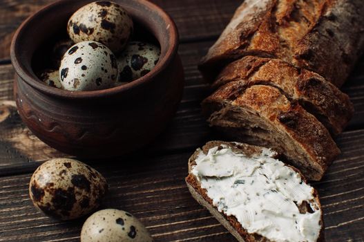 dark buckwheat bread is spread with cottage cheese with herbs in a cut on a wooden table near quail eggs in a clay plate in a rustic style. Snack and breakfast concept.