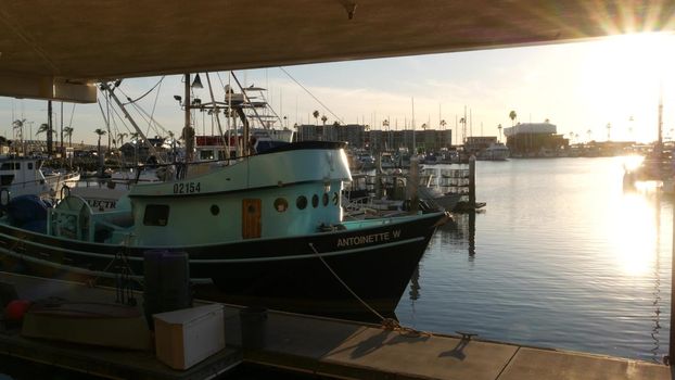 Oceanside, California USA - 26 Feb 2020: Harbor village with fisherman boats and yachts, pacific ocean coast marina, sea shore. Blue nautical vessel for fishing in port, fishery industry. Sunset light