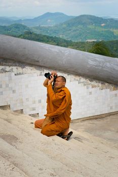 2019-11-06 / Phuket, Thailand - A buddhing monk in saffron robes kneels down to take a picture with a DSLR camera.