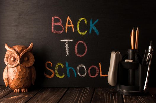 Back to school, the concept of parenting. An assortment of consumables, owl, notebook, pencils, stand for pens. The inscription on the slate black chalk. School supplies on a wooden table, rustic.