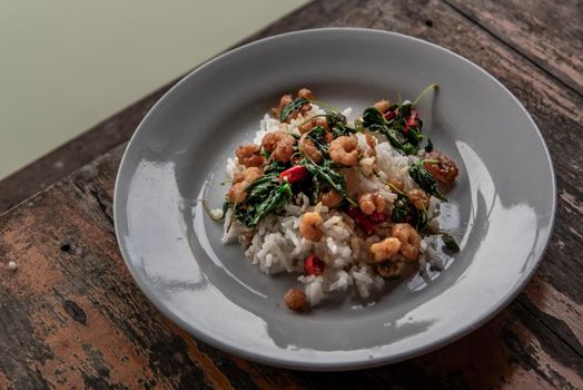 Rice topped with Stir fried thai basil with dwarf prawn served in white dish on wooden table, A world renowned delicacy from Thailand, Selective focus.