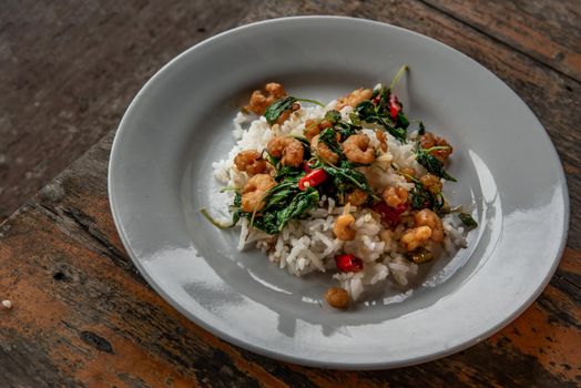 Rice topped with Stir fried thai basil with dwarf prawn served in white dish on wooden table, A world renowned delicacy from Thailand, Selective focus.