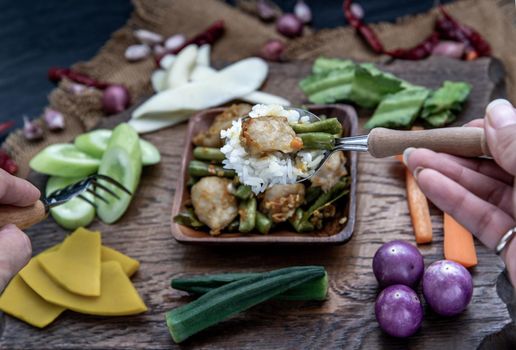 Thai food, The hand is using a spoon to scoop Stir Fried fish ball with Red Curry Paste and long bean with the ingredient and fresh vegetables on wood background.