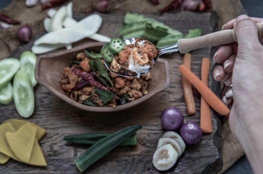 The hand is using a spoon to scoop Pork Crackling Chili Paste with fried kaffir lime leaves with the ingredient and fresh vegetables on wood background, view from above.