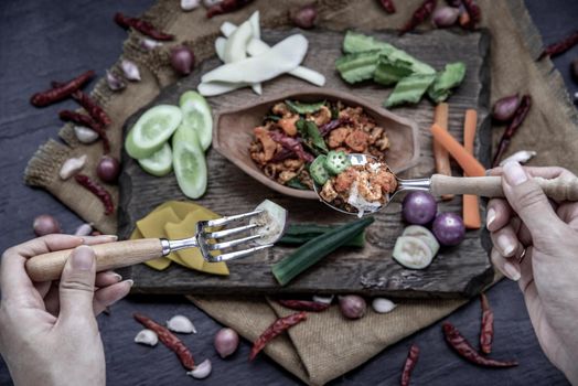 The hand is using a spoon to scoop Pork Crackling Chili Paste with fried kaffir lime leaves with the ingredient and fresh vegetables on wood background, view from above.