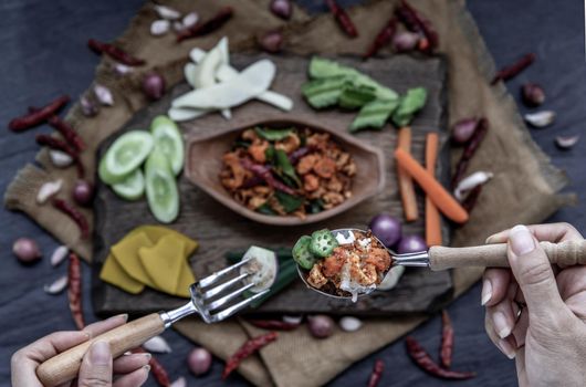 The hand is using a spoon to scoop Pork Crackling Chili Paste with fried kaffir lime leaves with the ingredient and fresh vegetables on wood background, view from above.