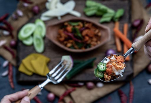 The hand is using a spoon to scoop Pork Crackling Chili Paste with fried kaffir lime leaves with the ingredient and fresh vegetables on wood background, view from above.