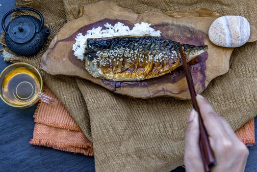 The hand is using chopsticks to pick up saba fish grilled on rice with teriyaki sauce on a wooden background served with hot tea, Japanese style.