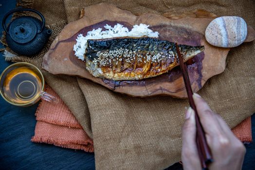 The hand is using chopsticks to pick up saba fish grilled on rice with teriyaki sauce on a wooden background served with hot tea, Japanese style.
