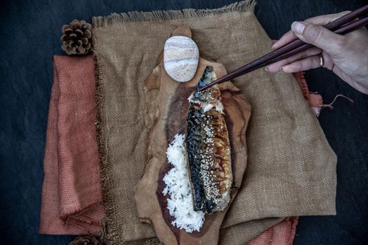 The hand is using chopsticks to pick up saba fish grilled on rice with teriyaki sauce on a sackcloth background, Japanese style.