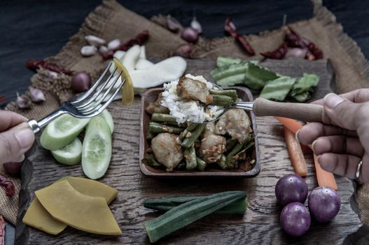 Thai food, The hand is using a spoon to scoop Stir Fried fish ball with Red Curry Paste and long bean with the ingredient and fresh vegetables on wood background.