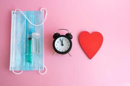 On a pink background-a mask, a syringe, a bottle of vaccine, a watch and a red wooden heart. The concept of medicine and health care.