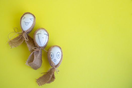 Three white eggs with funny faces in sackcloth on a yellow background. Easter Concept. Space for text.