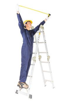 Young handyman in uniform standing on ladder while using tape measure on white, Cutout isolated on white background