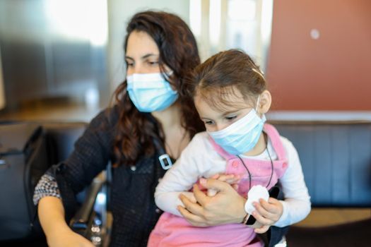 Portrait of woman lovingly hugging her young daughter while wearing face masks. Adorable view of little girl wearing pink overalls in the arms of her mother with blurry background. Life after Covid-19