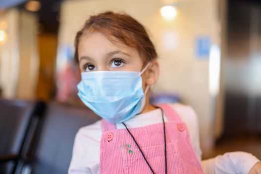 Portrait of adorable little girl wearing pink corduroy overalls and face mask. Close-up of beautiful young child in pink and white clothing with blurry background. New normal after Covid-19