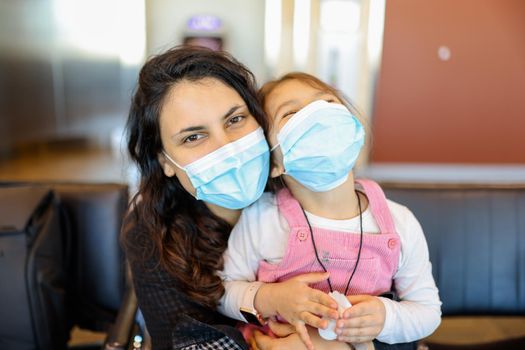 Portrait of woman lovingly hugging her young daughter while wearing face masks. Adorable view of little girl wearing pink overalls in the arms of her mother with blurry background. Life after Covid-19