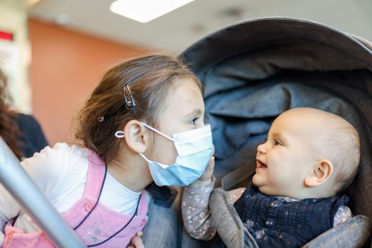 Portrait of adorable little girl wearing pink overalls and face mask next to her happy baby sister in stroller. Close-up of cute young children with blurry background. New normal after Covid-19