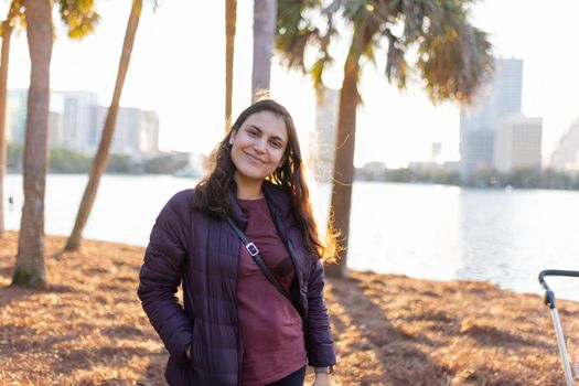 Portrait of beautiful woman with blurry palm trees and bright sunset as background. Smiling brunette woman in black jacket standing near a lake in the evening. Adventurous day outdoors