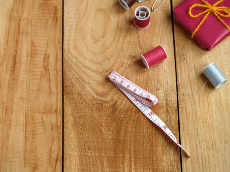 Tailor's measuring tape, thread coils and red gift box on brown wooden boards.