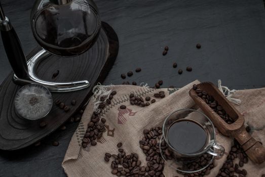 Coffee style, Cup of coffee and coffee beans roating with old wooden scoop and coffee beans around on the wooden and dark stone background. Oblique view from the top with copy space for your text.
