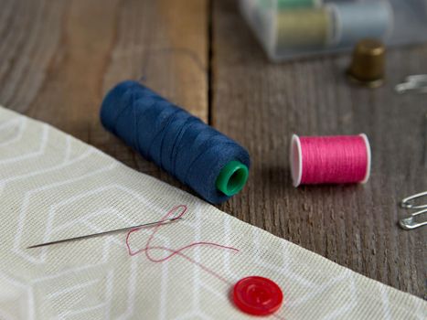 Coils with red and blue threads, needle with thread, gray fabric on brown wooden boards. Selective focus