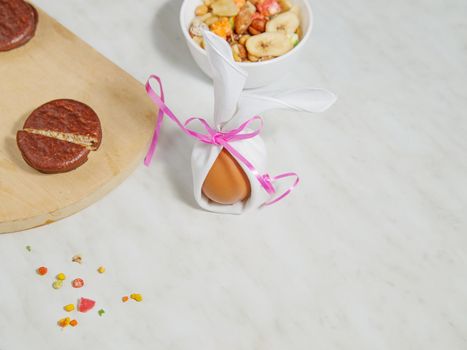 Chicken egg is wrapped in white material tied with red ribbon. Board of biscuits, jar of nuts on white table. Selective focus