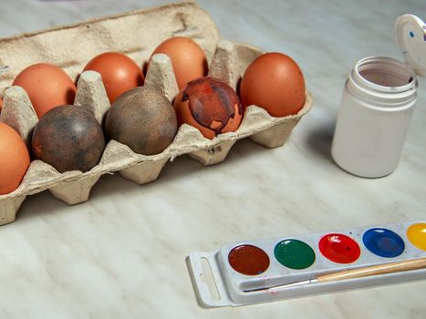 Painted chicken eggs in box next to brush, paint and jar on gray table.