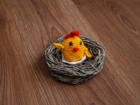 Yellow toy chicken in nest of gray woolen threads on brown surface.