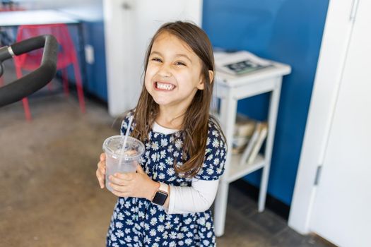 Adorable view of happy little girl wearing blue flowered dress and holding plastic cup in restaurant. Portrait of smiling young child with blue walls and white doors as background. Cute kids drinking