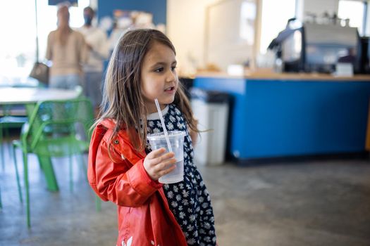 Adorable view of happy little girl wearing red jacket and blue flowered dress holding plastic cup in restaurant. Portrait of cute young child in cafe with blurry background. Lovely kids drinking