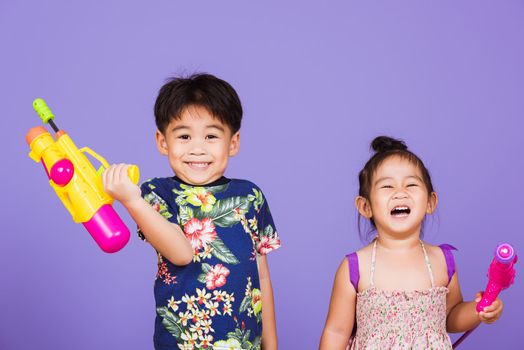 Two Happy Asian little boy and girl holding plastic water gun, Thai children funny hold toy water pistol and smile, studio shot isolated on purple background, Thailand Songkran festival day culture.