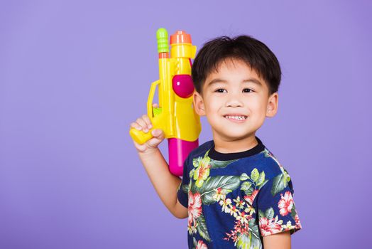 Thai kid funny hold toy water pistol and smiling, Happy Asian little boy holding plastic water gun, studio shot isolated on purple background, Thailand Songkran festival day national culture concept