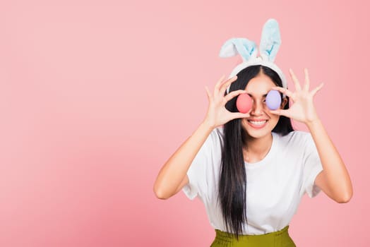 Happy beautiful young woman smiling wearing rabbit ears holding colorful Easter eggs front eyes, Thai female with bunny ear hold easter egg covering eye, studio shot isolated on pink background