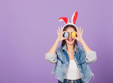 Happy beautiful young woman smiling wearing rabbit ears and denims holding colorful Easter eggs front eyes, Thai female with bunny ear, easter egg cover eye, studio shot isolated on purple background